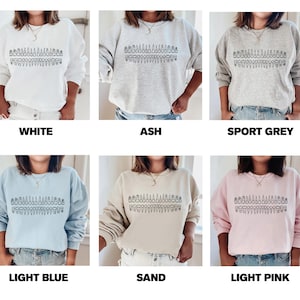 May include: A variety of colors of sweatshirts with a graphic of a row of trees on the front. The colors are white, ash, sport grey, light blue, sand, and light pink.