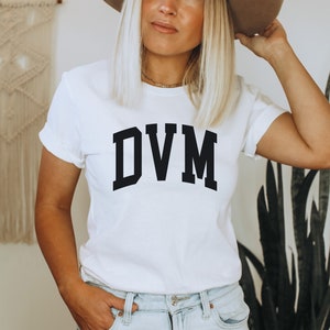 May include: White t-shirt with black text that reads "DVM" in a collegiate font.