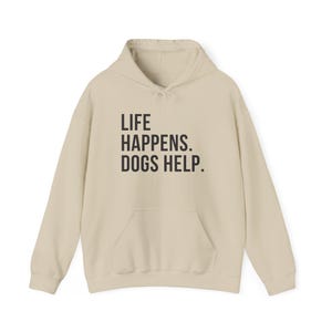 May include: A cream-colored hoodie with a front pocket and drawstring hood. The front of the hoodie features the text "LIFE HAPPENS. DOGS HELP." in a bold, black font. A casual and comfortable apparel item.
