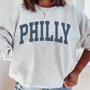 Könnte beinhalten: Grauer Pullover mit dem Wort "PHILLY" in dunkelblauen Buchstaben.