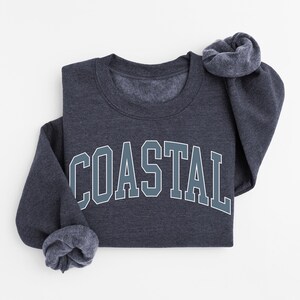May include: A dark gray sweatshirt with the word "COASTAL" in large, block letters across the front. The letters are outlined in white and filled with a teal color. The sleeves are folded up, and the sweatshirt is on a white background.