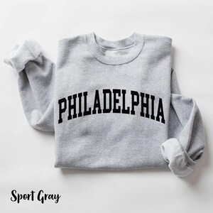 May include: A gray sweatshirt with the word "PHILADELPHIA" printed in black on the front. The sweatshirt is a classic crew neck style.