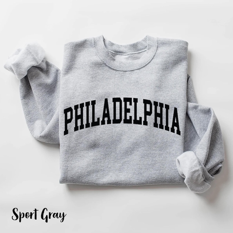 May include: A gray sweatshirt with the word "PHILADELPHIA" printed in black on the front. The sweatshirt is a classic crew neck style.