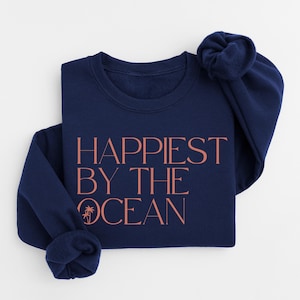May include: A navy blue sweatshirt with the text "Happiest by the Ocean" in a coral color. The design includes a small palm tree graphic. The sweatshirt is folded neatly, with one sleeve bunched up.