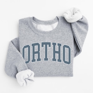 May include: A heather gray sweatshirt with the word "ORTHO" in a blue collegiate font across the chest. The sleeves are cuffed, revealing a white interior. The sweatshirt is folded neatly.
