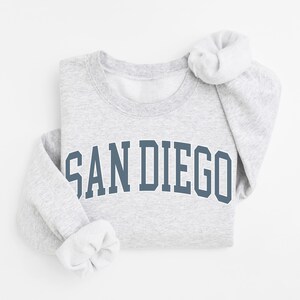 May include: A light gray sweatshirt with the text "SAN DIEGO" in a blue, arched font. The sleeves are folded up, revealing a white interior. The sweatshirt is folded neatly on a white surface.