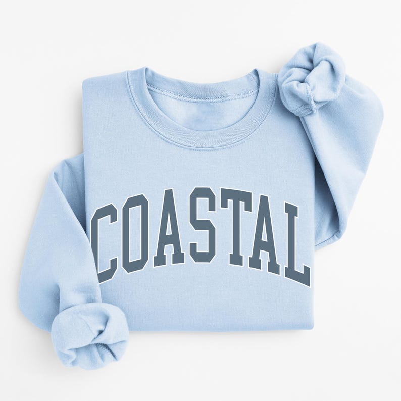May include: A light blue sweatshirt with the word "COASTAL" printed in dark gray block letters across the chest. The sleeves are folded, and the crew neck sweatshirt is on a white background.