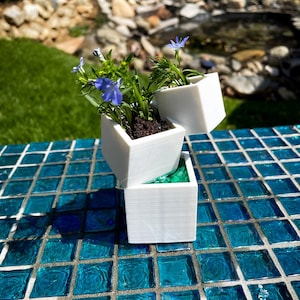 May include: Three white square planters stacked on top of each other. The top planter has a small blue flower plant, the middle planter has a larger blue flower plant, and the bottom planter is empty. The planters are sitting on a blue tiled surface.