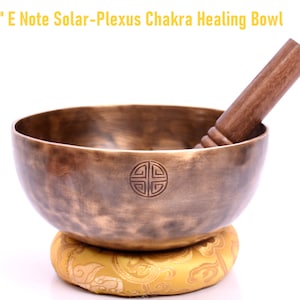Puede incluir: Un cuenco curativo del chakra del plexo solar E Note de 20 cm. El cuenco de color bronce tiene un emblema decorativo y descansa sobre un cojín amarillo. Una maza de madera descansa dentro del cuenco. El texto en la imagen dice: 8" E Note Solar-Plexus Chakra Healing Bowl.
