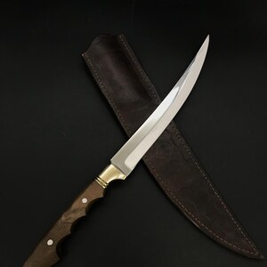 May include: A long, curved, silver knife with a brown wooden handle and a leather sheath. The knife is lying on a black surface.