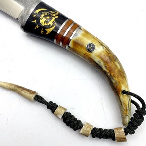 May include: A black-handled knife with a wolf design on the handle. The handle is made of bone and has a brown and gold inlay. The knife has a black cord lanyard with bone beads.