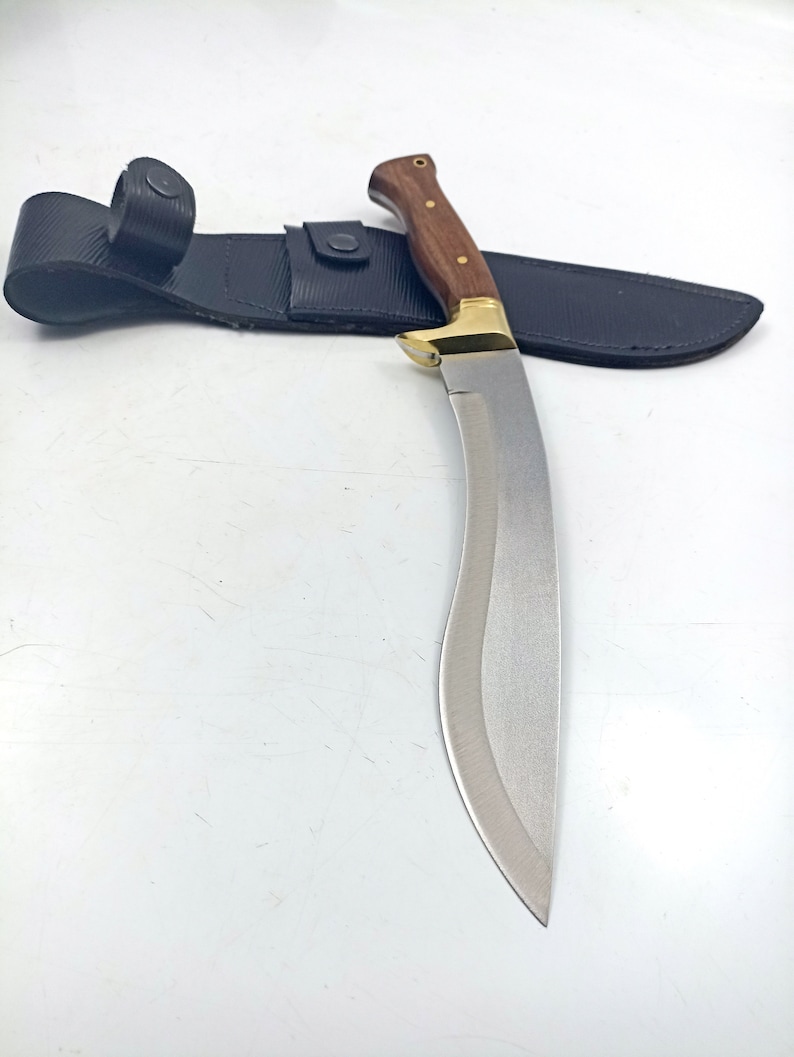 Puede incluir: Una hoja de plata curva con un mango de madera y una funda de cuero negro. La hoja es afilada y puntiaguda, y el mango est&aacute; decorado con detalles de lat&oacute;n.