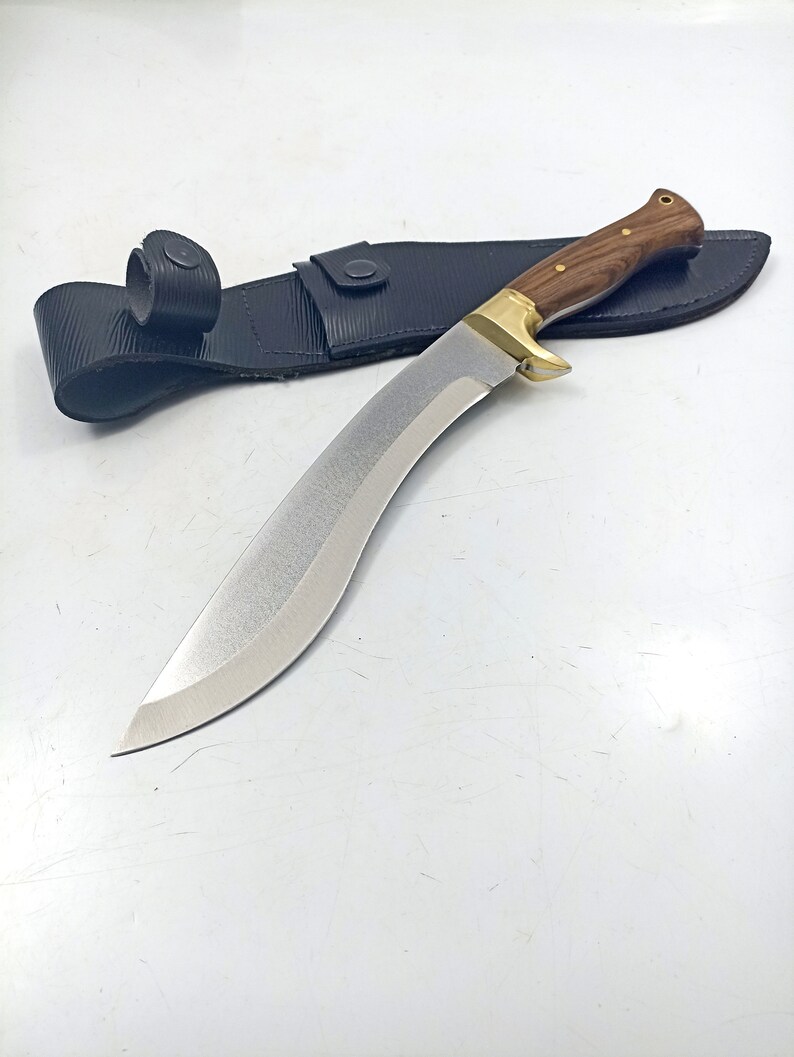 Puede incluir: Un cuchillo curvo de color plateado con mango de madera y funda de cuero negro. El cuchillo tiene una punta afilada y un borde dentado.