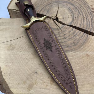 May include: A brown leather sheath with gold stitching and a decorative design. The sheath is for a dagger with a wooden handle.