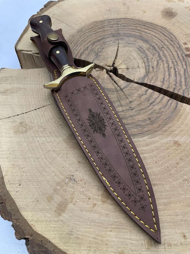 May include: A brown leather sheath with gold stitching and a decorative design. The sheath is for a dagger with a wooden handle.