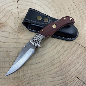 May include: A folding pocket knife with a wooden handle and a decorative metal bolster. The blade is silver and the handle is a dark brown color. The knife comes with a black leather sheath with brass-colored rivets.