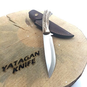 May include: A hunting knife with a polished steel blade and a handle made of light brown and cream-coloured material. The knife rests on a wooden surface with the text "YATAGAN KNIFE" burned into it. A brown leather sheath is behind the knife.