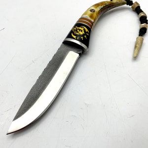 May include: A small, curved knife with a brown and white antler handle. The blade is made of steel and has a textured finish. The handle is decorated with a gold wolf design. The knife has a lanyard attached to the handle.