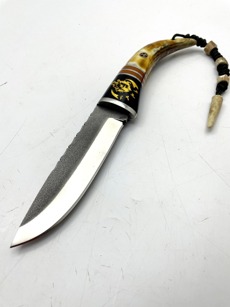 May include: A small, curved knife with a brown and white antler handle. The blade is made of steel and has a textured finish. The handle is decorated with a gold wolf design. The knife has a lanyard attached to the handle.