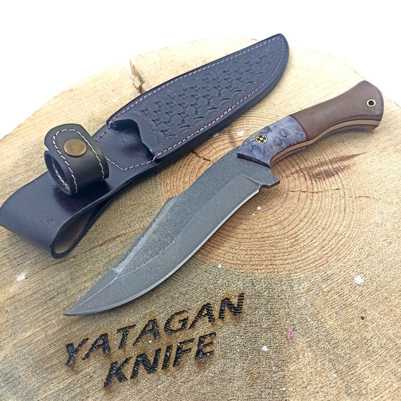 May include: A black and gray knife with a wooden handle and a leather sheath. The knife is on a wooden surface with the text "YATAGAN KNIFE" burned into the wood.