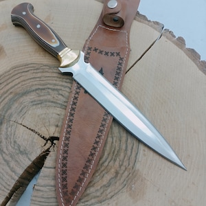 Op de afbeelding: Een bruine leren schede met decoratieve stiksels en een zilveren metalen knoop. Een zilveren en gouden mes met een bruin en wit handvat zit in de schede.