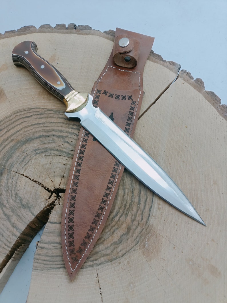Op de afbeelding: Een bruine leren schede met decoratieve stiksels en een zilveren metalen knoop. Een zilveren en gouden mes met een bruin en wit handvat zit in de schede.