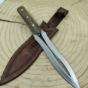 Peut inclure: Un couteau artisanal avec un manche en bois et une lame à double tranchant. Le couteau est partiellement gainé dans un étui en cuir marron avec des coutures décoratives. La poignée comporte trois rivets argentés.