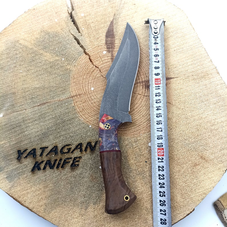 May include: A YATAGAN KNIFE with a dark gray blade and a brown wooden handle with a blue and red pattern. The knife is on a wooden surface with a measuring tape beside it.