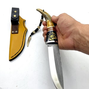 May include: A hunting knife with a brown antler handle and a black leather sheath. The blade is made of Damascus steel and has a decorative pattern.