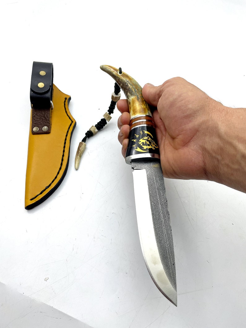 May include: A hunting knife with a brown antler handle and a black leather sheath. The blade is made of Damascus steel and has a decorative pattern.