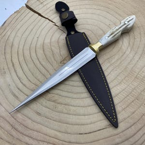May include: A brown leather sheath with a yellow stitch detail holds a silver knife with a white bone handle and a gold-colored metal accent. The knife has a pointed tip and a straight blade.