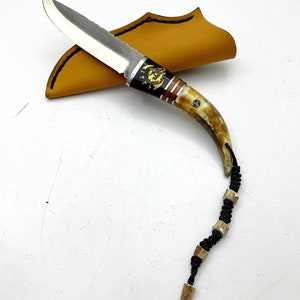 May include: A small, black-handled knife with a silver blade and a decorative antler handle. The knife is in a brown leather sheath and has a black cord with antler beads.