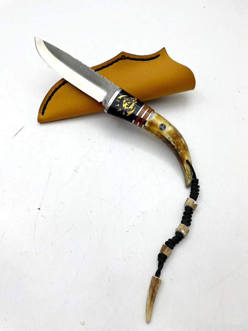 May include: A small, black-handled knife with a silver blade and a decorative antler handle. The knife is in a brown leather sheath and has a black cord with antler beads.