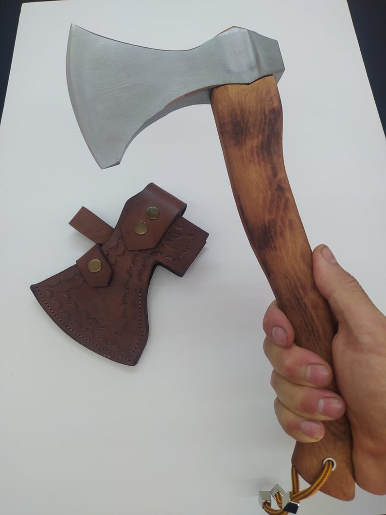 May include: Axe with a silver blade and a wooden handle. The handle is a light brown color. A brown leather sheath with a decorative pattern and two brass-colored snaps is included.