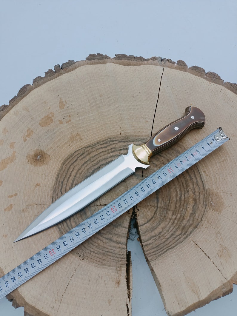 Op de afbeelding: Een zilveren dolk met een bruin houten handvat en een messing accent. De dolk ligt op een houten oppervlak met een meetlint ernaast. De tape meet 94 cm.