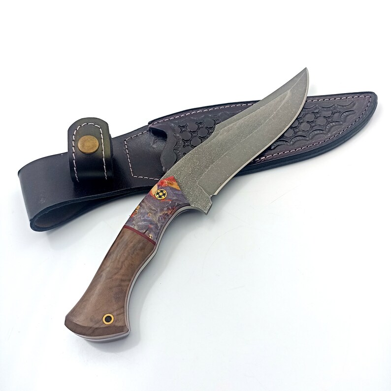 May include: A brown leather sheath with a decorative pattern holds a hunting knife with a wood handle and a patterned steel blade.