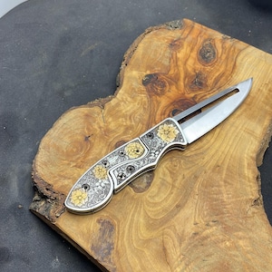 Peut inclure: Un couteau pliant argent&eacute; avec un motif floral. Le manche pr&eacute;sente des fleurs jaunes et argent&eacute;es et des d&eacute;tails complexes. La lame est ouverte, r&eacute;v&eacute;lant un tranchant. Le couteau repose sur une surface en bois.
