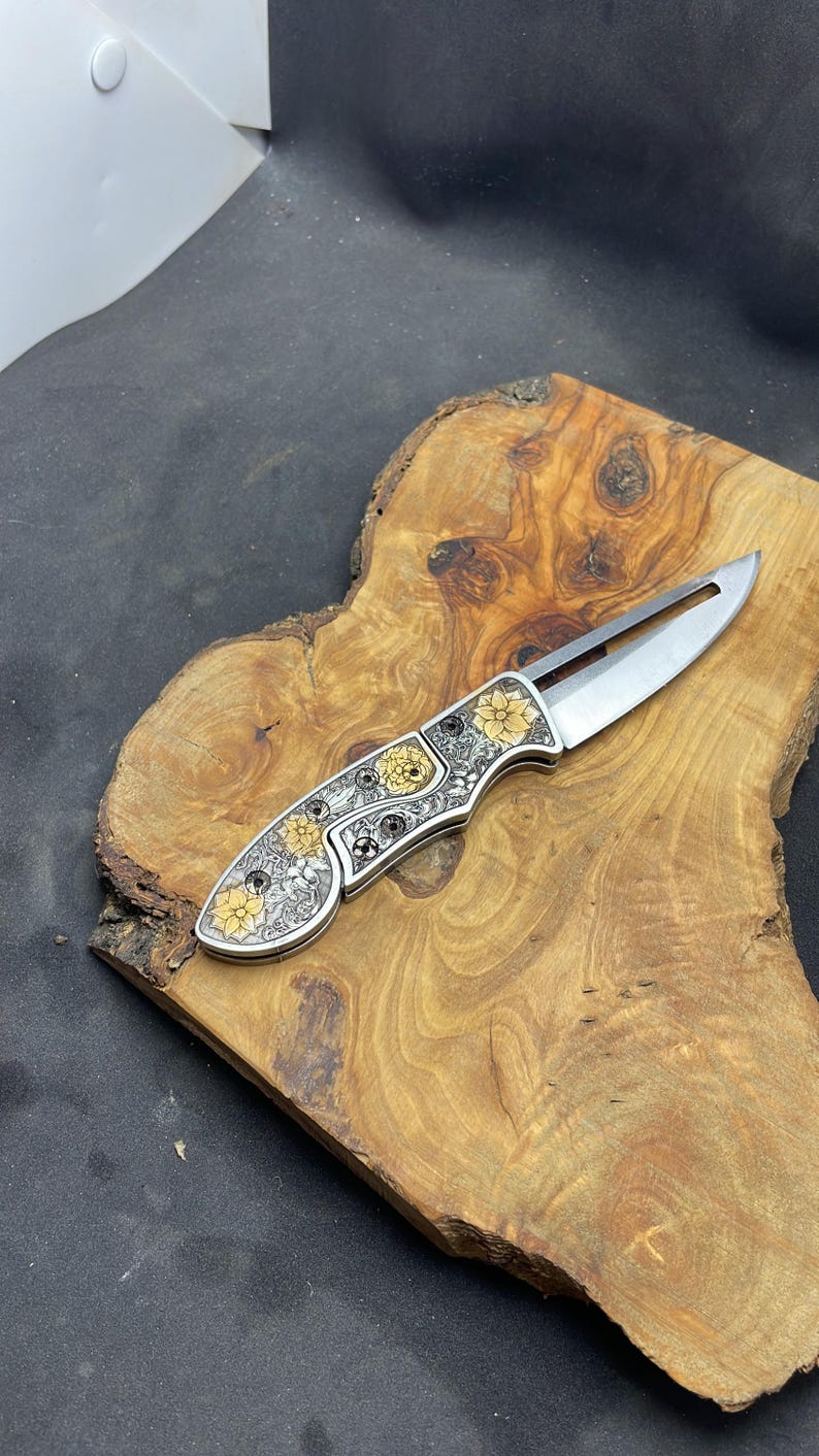 Peut inclure: Un couteau pliant argent&eacute; avec un motif floral. Le manche pr&eacute;sente des fleurs jaunes et argent&eacute;es et des d&eacute;tails complexes. La lame est ouverte, r&eacute;v&eacute;lant un tranchant. Le couteau repose sur une surface en bois.