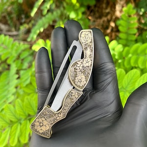 Peut inclure: Un couteau pliant avec une lame argent&eacute;e et un manche d&eacute;coratif. Le manche est de couleur dor&eacute;e et pr&eacute;sente un motif floral grav&eacute;. Le couteau est tenu dans une main gant&eacute;e de noir sur un fond vert flou.