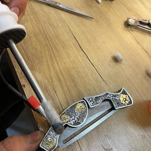 Peut inclure: Gros plan d&eacute;taill&eacute; d'un couteau de poche argent&eacute; et dor&eacute; avec des motifs floraux. Le couteau est travaill&eacute; avec un outil, mettant en valeur le savoir-faire complexe. La lame est partiellement ouverte, r&eacute;v&eacute;lant son tranchant.