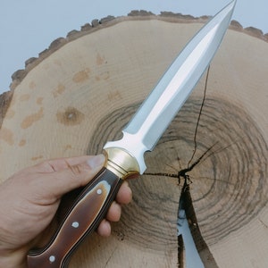 Op de afbeelding: Een zilveren dolk met een bruin en zwart houten handvat. Het handvat heeft een gouden metalen accent aan de bovenkant. De dolk ligt op een houten oppervlak.