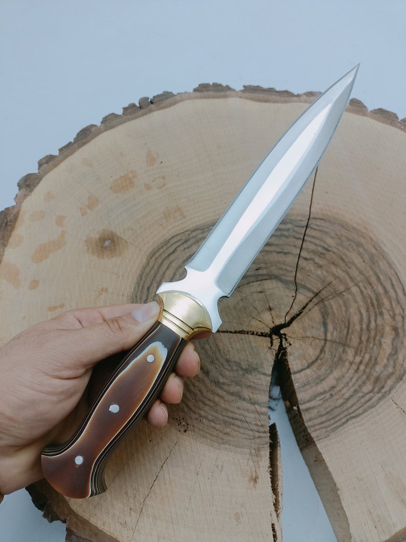 Op de afbeelding: Een zilveren dolk met een bruin en zwart houten handvat. Het handvat heeft een gouden metalen accent aan de bovenkant. De dolk ligt op een houten oppervlak.