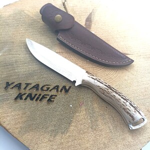 May include: A hunting knife with a stainless steel blade and a handle with a light brown, speckled pattern. The knife is accompanied by a brown leather sheath. The text "YATAGAN KNIFE" is carved into the wooden surface.