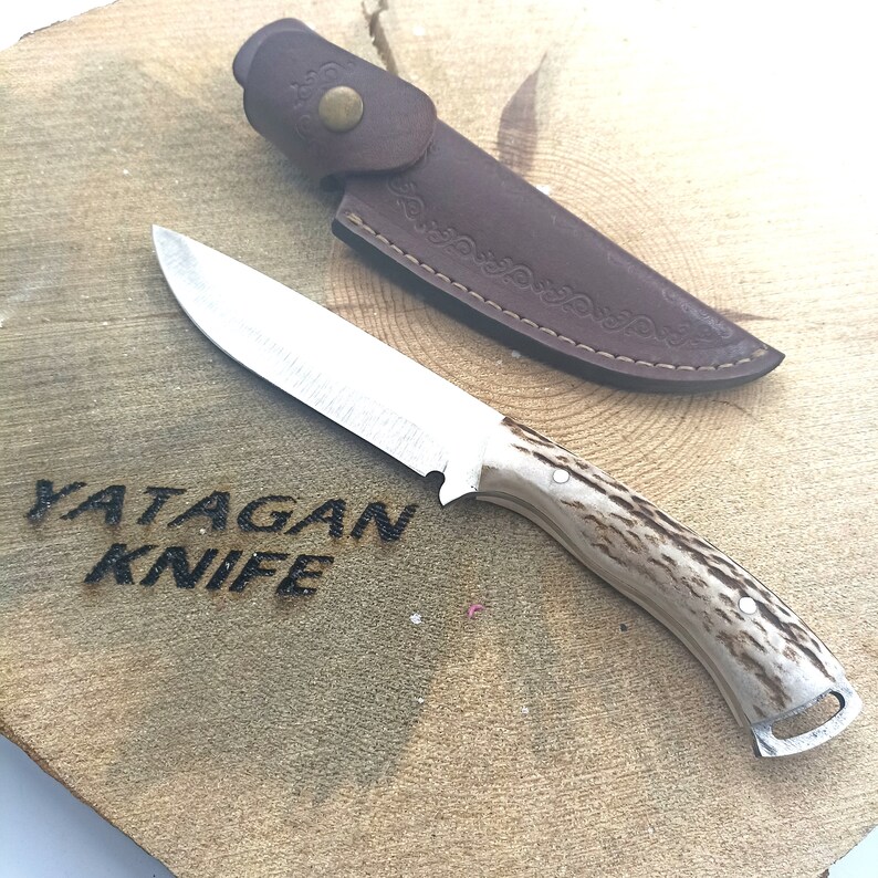 May include: A hunting knife with a stainless steel blade and a handle with a light brown, speckled pattern. The knife is accompanied by a brown leather sheath. The text "YATAGAN KNIFE" is carved into the wooden surface.