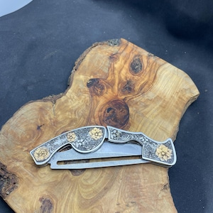 Peut inclure: Un couteau pliant argent&eacute; avec des gravures florales dor&eacute;es. Le couteau est ouvert, r&eacute;v&eacute;lant la lame. La poign&eacute;e pr&eacute;sente des motifs floraux d&eacute;taill&eacute;s. Le couteau repose sur une surface en bois avec un grain naturel.