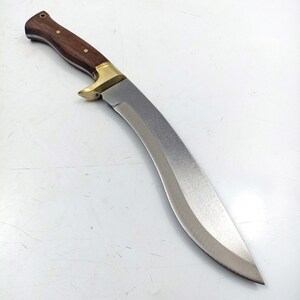 Puede incluir: Un cuchillo curvo de color plateado con un mango de madera. El mango es marr&oacute;n con un acento de lat&oacute;n.