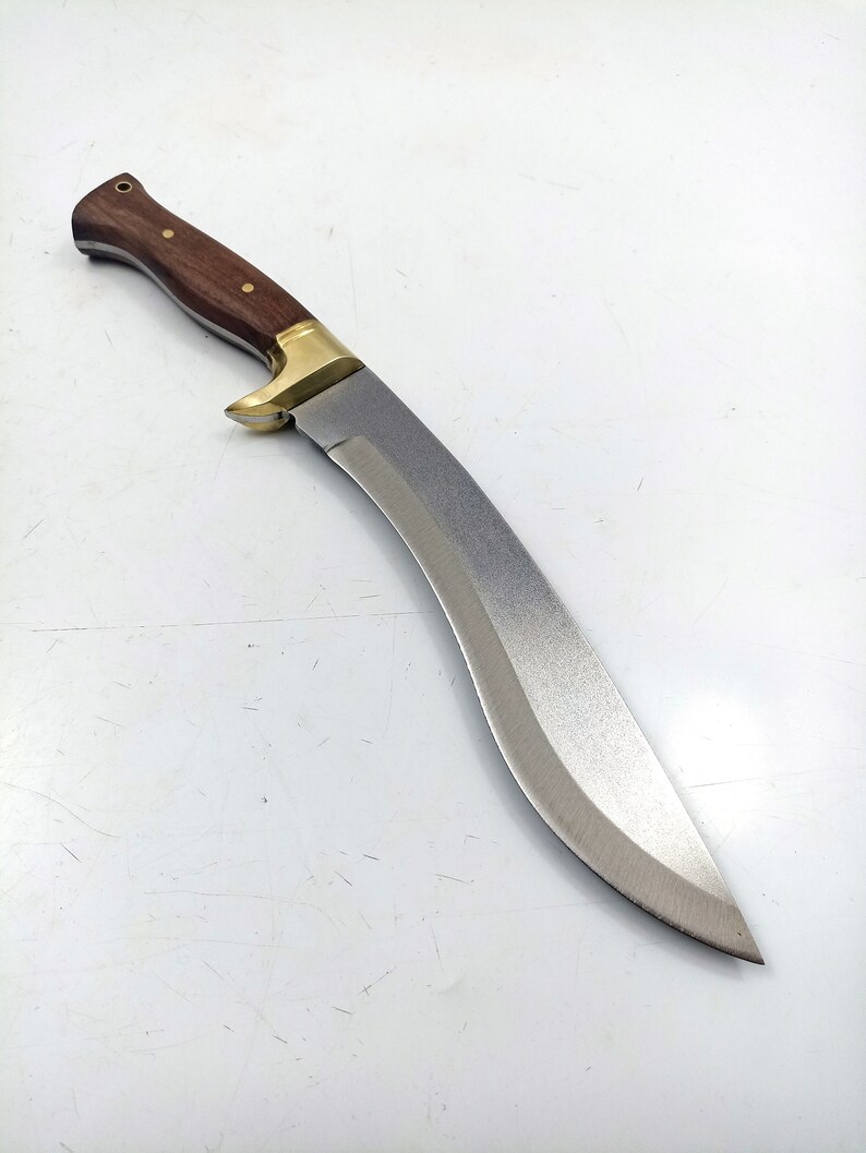 Puede incluir: Un cuchillo curvo de color plateado con un mango de madera. El mango es marr&oacute;n con un acento de lat&oacute;n.