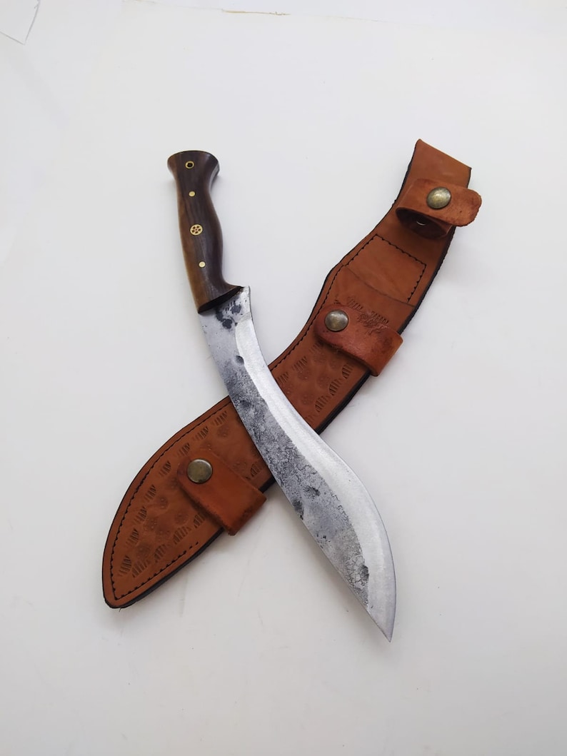 May include: A curved, silver-colored knife with a brown wooden handle and a brown leather sheath. The sheath has decorative stitching and a brass button.