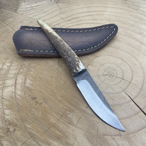 May include: A small, handmade knife with a brown leather sheath. The knife has a  handle made of antler and a  blade with a  slight curve.