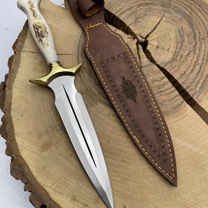 May include: A silver dagger with a white antler handle and a gold-coloured guard. The dagger is on a wooden surface and has a brown leather sheath with decorative stitching.
